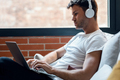 Smart man working with her laptop while listening music with headphones sitting on a couch at home - PhotoDune Item for Sale