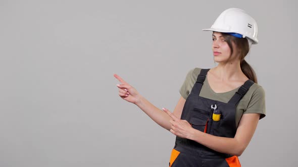 Portrait of Attractive Young Builder Woman in Helmet and Jumpsuit Smiling and Pointing Finger at