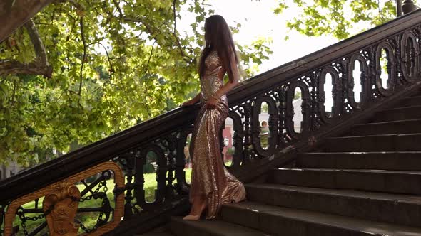 Attractive Young Fit Woman in a Long Golden Shiny Dress Walks Down on the Stairs on Highheels in