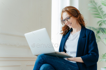 Professional female employer enjoys working process, sits crossed legs with laptop device