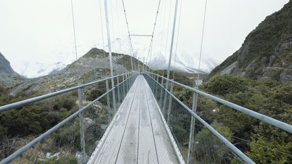 Walking across a swing bridge over a running lake surrounded by snow capped mountains and peaks duri