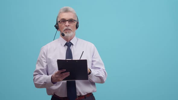 An adult male call centre with headphones is talking and holding a paper tablet in blue background