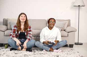 a joystick. Black girl wearing eyeglasses and beige sweater, caucasian girl wearing plaid shirt. Girls playing games together.