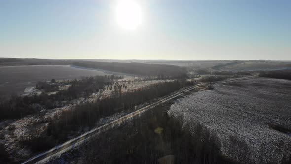 Flying over the road among the fields and woods towards the low sun