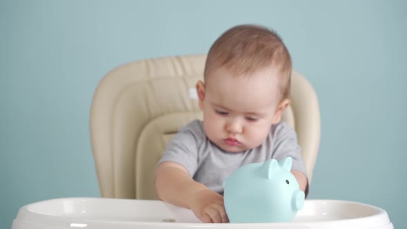 Baby 12-17 months throws a Russian ruble coin into a blue piggy bank sitting on a chair