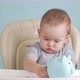 Baby 12-17 months throws a Russian ruble coin into a blue piggy bank sitting on a chair - VideoHive Item for Sale