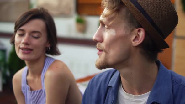 Young Carefree Couple Sitting Outdoors and Man in a Hat Smoking Marijuana Joint Smiling Slow Motion