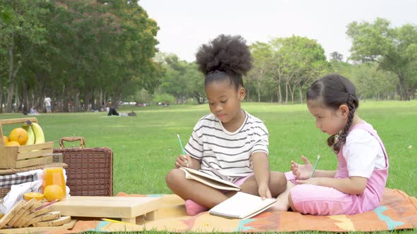 Two little girl writing homework on notebook together while sitting and lying on mat in the park