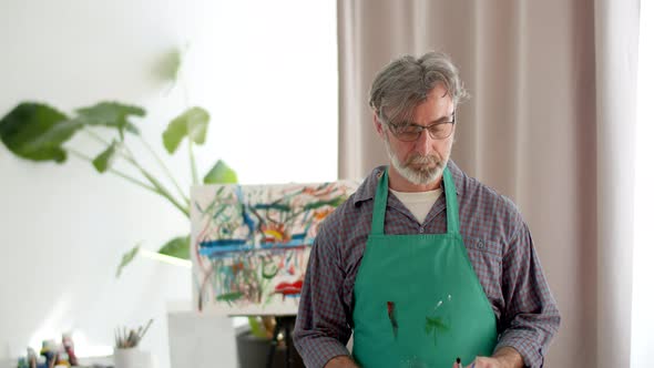 Elderly Male Artist Master in an Apron Stands in Studio Holding Brushes with Arms Crossed on Chest