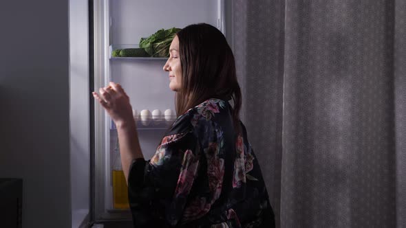 Silhouette of Woman Eating Eclair at Fridge in Dark Kitchen