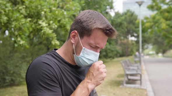 A Young Man in a Face Mask Feels Sick, Coughs and Has a Headache As He Sits on a Bench in a Park