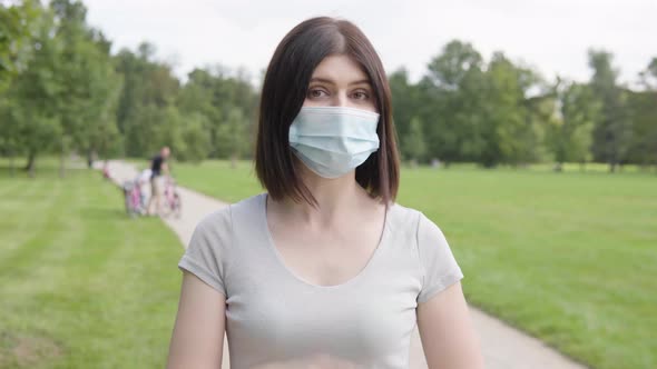 A Young Caucasian Woman in a Face Mask Talks to the Camera on a Pathway in a Park on a Sunny Day