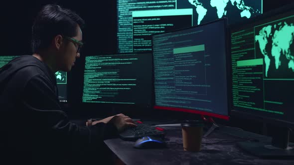 Asian Male Hacker Hacking With Multiple Computer Screens In Dark Room