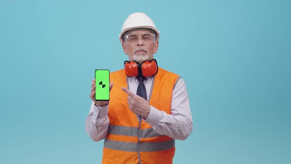 Working Engineer Man in a Protective Uniform Holds a Phone with an App on Blue Studio Background