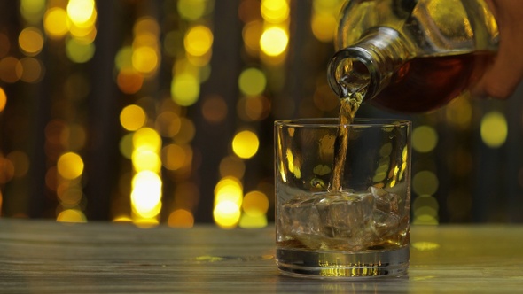 Pouring of Golden Whiskey, Cognac or Brandy From Bottle Into Glass with Ice Cubes. Shiny Background