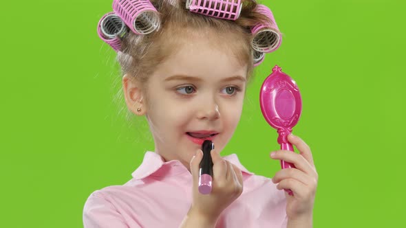 Child in Curlers Holds a Mirror in Her Hands and Paints Lipstick with Lipstick. Green Screen