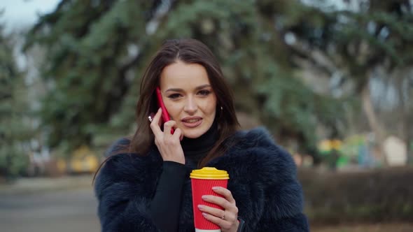 Girl in fur coat with a phone and coffee outdoors