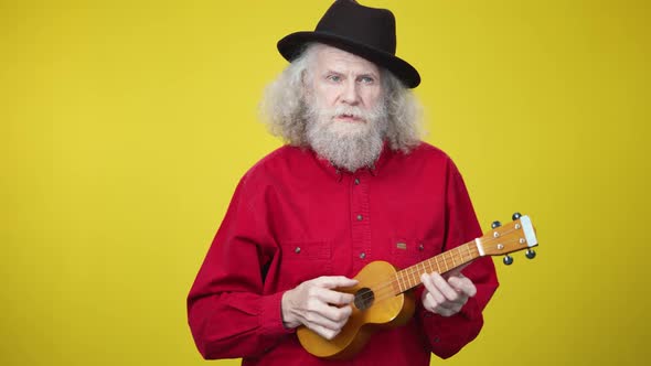 Elegant Senior Man in Red Shirt and Black Hat Singing and Playing Ukulele at Yellow Background