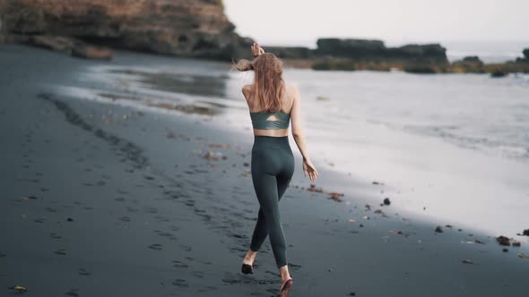 Backside View Woman Walks at Black Beach Raises Her Arms to High Turns Around