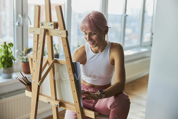 Smiling woman with pink hair draws on canvas at wooden easel on balcony