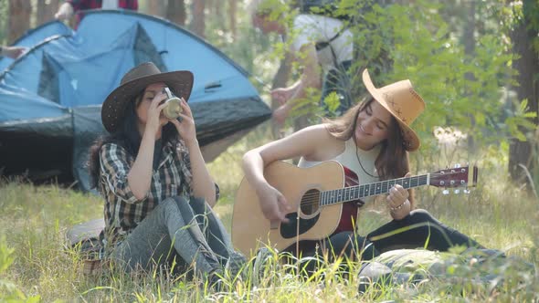 Beautiful Young Women Singing and Playing Ukulele As Men Putting Up Tent at the Background. Portrait