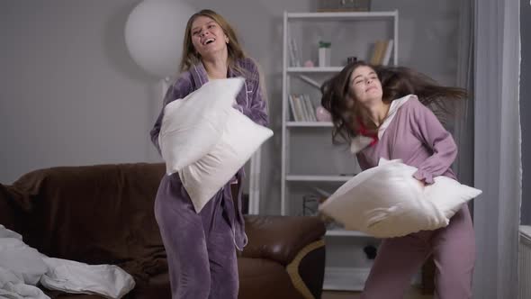 Two Joyful Caucasian Young Women Dancing with Pillows at Pajama Party Indoors