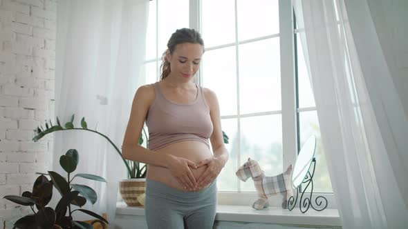 Closeup Joyful Pregnant Woman Making Heart Hand on Naked Belly at Home.