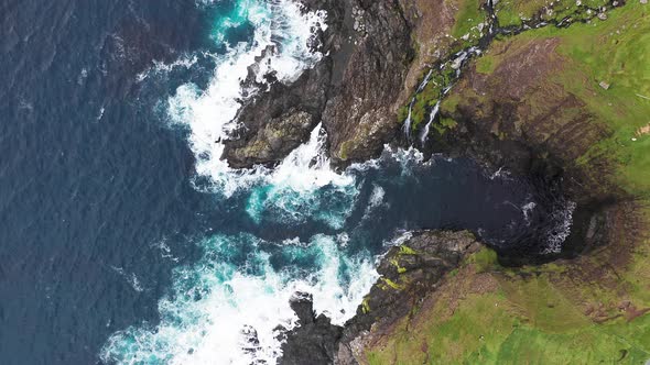 Aerial View of Waves Break on Rocks of Faroe Islands Cliffs in a Blue Ocean