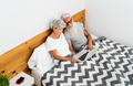 Happy senior couple having fun using computer while lying in bed below blankets - PhotoDune Item for Sale
