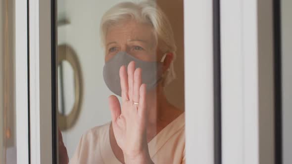 Senior caucasian woman wearing face mask looking out of window at home