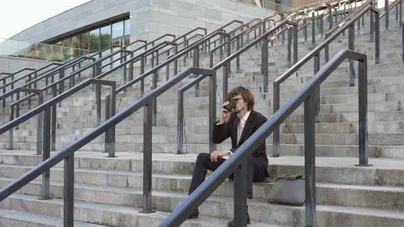 A businessman on a lunch break drinks coffee while sitting on the steps of the stairs.