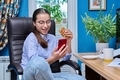 Teenage girl sitting on armchair near computer, looking in smartphone, eating snack - PhotoDune Item for Sale