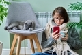 Woman at home using smartphone, warming with cat near heating radiator - PhotoDune Item for Sale
