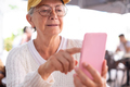 Close-up on senior woman with hat and eyeglasses using phone while sitting at a cafe table - PhotoDune Item for Sale
