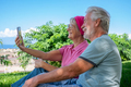 Portrait of senior smiling couple sitting on the grass in public park taking selfie with phon - PhotoDune Item for Sale
