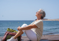 Happy senior woman sitting near the beach with her face facing the sun - PhotoDune Item for Sale