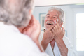 Skin care. Handsome senior man applying cream at his face and looking at himself in the mirror - PhotoDune Item for Sale