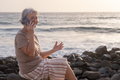Beautiful senior woman sitting at the beach at sunset talking on smart phone smiling - PhotoDune Item for Sale