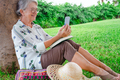 Smiling senior woman sitting in the meadow of public park against a tree trunk looking at phone - PhotoDune Item for Sale