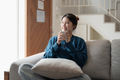 Happy young asian woman sitting on a couch with a cup of coffee at home - PhotoDune Item for Sale