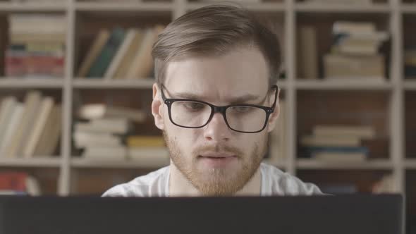 Close-up of Focused Young Man in Eyeglasses Looking at Laptop Screen. Portrait of Caucasian Brunette