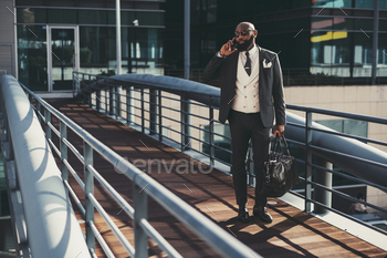 king on the phone while passing on an overhead passage; an African bearded businessman is phoning while standing on the pedestrian bridge with his bag