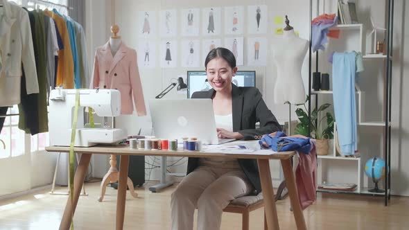 Smiling Asian Female Designer In Business Suit Working On A Laptop While Designing Clothes