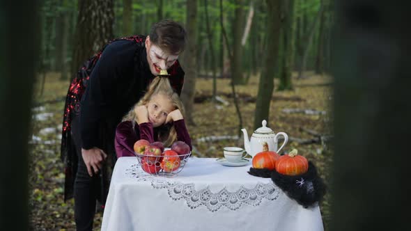 Portrait of Bored Caucasian Beautiful Girl Sitting at Table in Forest As Man in Vampire Costume and