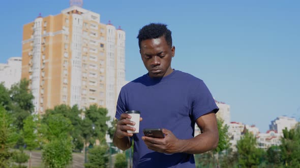 Afroamerican Man Uses Mobile Phone Holding White Cup with Coffee or Tea in Hand