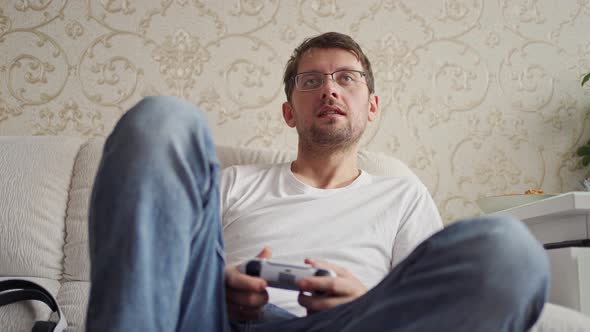 a Young Man Sits on the Couch and Enthusiastically Plays a Console Holds a Joystick in His Hands