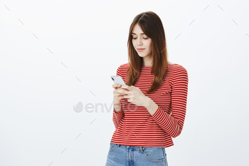 -looking woman in casual outfit, holding smartphone and typing message while standing half-turned over gray background, using gadget for playing games. Technology concept