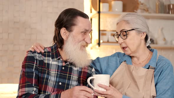 Caring Senior Woman Giving Tea To Senior Man in the Home Kitchen