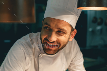 Close up of smiling male chef in uniform standing on kitchen of restaurant and looking