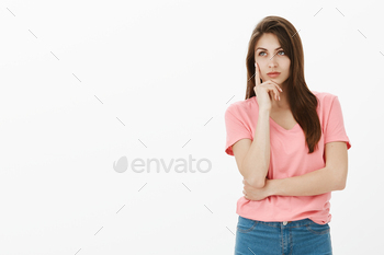 ing mean. Dreamy attractive and confident young woman in pink t-shirt, holding palm on cheek and gazing at upper left corner with slight smile.